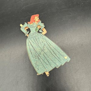 Puede incluir: Recorte de cart&oacute;n de estilo vintage de una mujer con un vestido verde azulado con un estampado floral. La mujer tiene el pelo rojo y posa con las manos en las caderas. El recorte tiene un borde dorado.