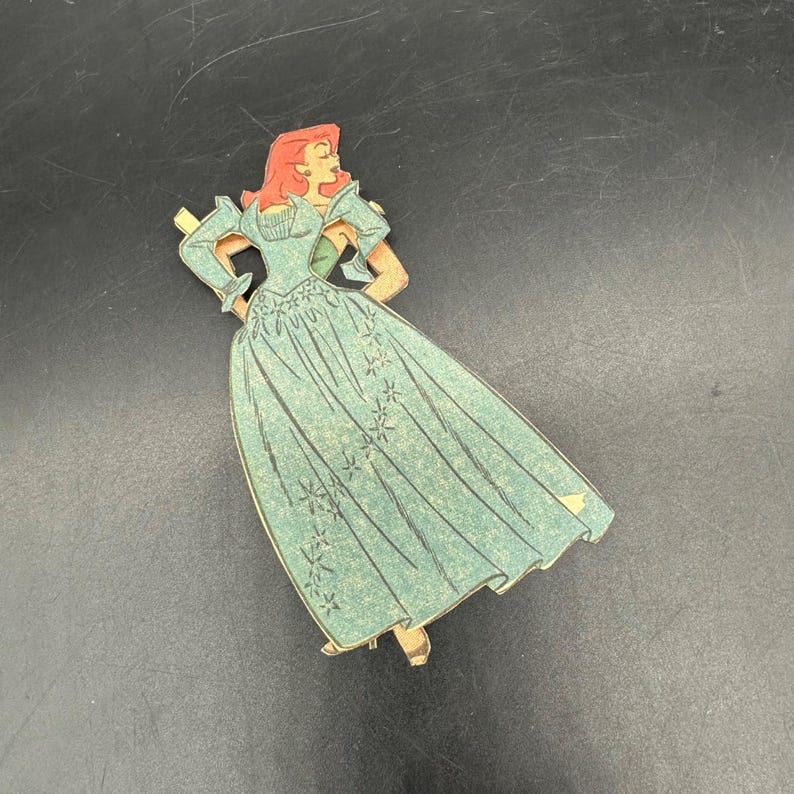 Puede incluir: Recorte de cart&oacute;n de estilo vintage de una mujer con un vestido verde azulado con un estampado floral. La mujer tiene el pelo rojo y posa con las manos en las caderas. El recorte tiene un borde dorado.