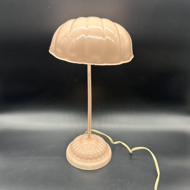 K&ouml;nnte beinhalten: Eine Vintage-Tischlampe in Hellrosa mit einem gewellten Lampenschirm. Die Lampe hat einen schlanken Stiel und einen runden, dekorativen Sockel. Das Stromkabel ist cremefarben. Die Lampe hat eine weiche, neutrale Farbe.