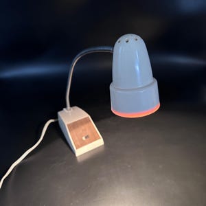 May include: A vintage desk lamp with a white shade and a flexible, silver-colored neck. The base is rectangular with a wood-grain panel. The lamp emits a warm, orange glow from the bottom of the shade. The lamp is plugged into a white cord.