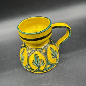 May include: A handcrafted yellow ceramic pitcher with a unique design. The pitcher features a rounded base with a decorative pattern in yellow, green, and black. The top section has a black stripe and a green rim, with a matching handle.
