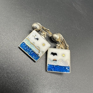 May include: A pair of silver clip-on earrings with square-shaped blue, white and gold mosaic designs. The mosaic designs feature a white background with blue and gold accents, and a black bird silhouette.
