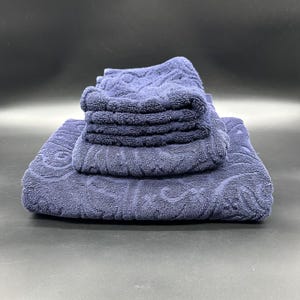May include: A set of navy blue bath towels, neatly stacked. The arrangement includes a large towel at the base, a medium-sized towel on top, and a pile of smaller face cloths. The towels feature a textured, patterned design.