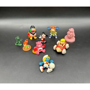 May include: A collection of colorful vintage figurines, including a penguin, Mickey Mouse, a Care Bear, a Smurf, and other cartoon characters. The figurines are made of plastic and are arranged on a dark surface.