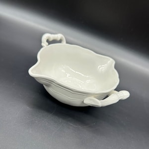 Puede incluir: Tazón de servir de porcelana blanca con dos asas ornamentadas. El tazón tiene una forma redondeada y un acabado liso y brillante.