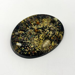 May include: An oval-shaped black resin piece embedded with gold glitter, small multicolored beads, and various smooth stones. The surface is glossy and smooth, featuring a mix of metallic gold flakes and tiny circular accents throughout the dark base.