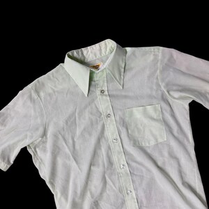 May include: A light green short-sleeved button-down shirt with a pointed collar and a chest pocket. The shirt has silver-colored buttons and a relaxed fit. The fabric appears to be lightweight and slightly textured, suitable for warmer weather.