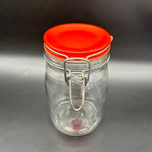 Könnte beinhalten: Ein klares Glasgefäß mit einem roten Deckel und einem Metallverschluss. Das Gefäß ist leer.