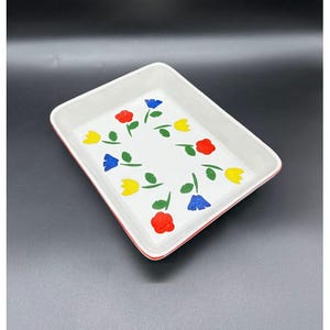 May include: Rectangular white ceramic baking dish with a red rim. The interior features a floral pattern of red, yellow, and blue flowers with green stems. The dish is set against a dark background.