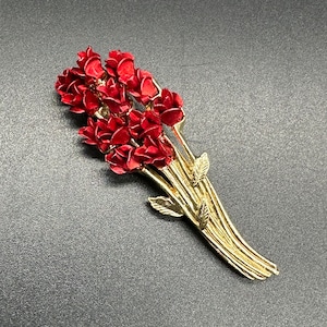 May include: A gold-toned brooch with a bouquet of red roses. The roses are clustered together and have a slightly textured finish. The stems are gold-toned and have small leaves.