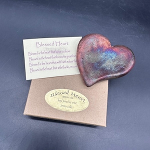 May include: A heart-shaped sculpture with a metallic finish in shades of purple, blue, and gold. The sculpture is next to a card with the text "Blessed Heart" and a box with a sticker that reads "Blessed Heart, hand formed by artist Jeremy Diller."