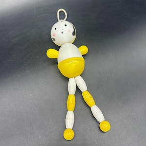 May include: A vintage toy doll with a white head and body, yellow arms and a yellow skirt. The doll's head has black and white details, with a loop at the top. The legs are made of white and yellow beads. The doll is on a dark background.