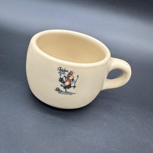 May include: A beige ceramic coffee mug with a cartoon rooster holding a golf club. The mug has the text "Chicken in the Rough" and "Reg. U.S. Pat. Off."