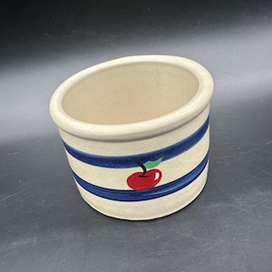 May include: A beige ceramic crock with blue horizontal stripes and a red apple design. The crock has a slightly angled rim and is suitable for kitchen or home decor. The apple has a green leaf.