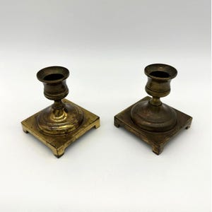 Puede incluir: Un par de candelabros de latón a juego con una base cuadrada de cuatro patas pequeñas y un tallo cilíndrico central que se ensancha en la parte superior para formar una copa circular para sostener una vela. El metal tiene un acabado pulido.