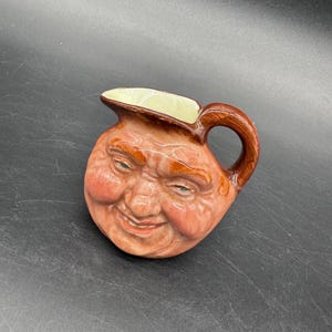 Puede incluir: Una jarra de cerámica con un diseño de cara sonriente. La jarra tiene un asa y un pico marrones, con un interior color crema. La cara está detallada con mejillas rojas, cejas naranjas y ojos azules. El color general es marrón rojizo.