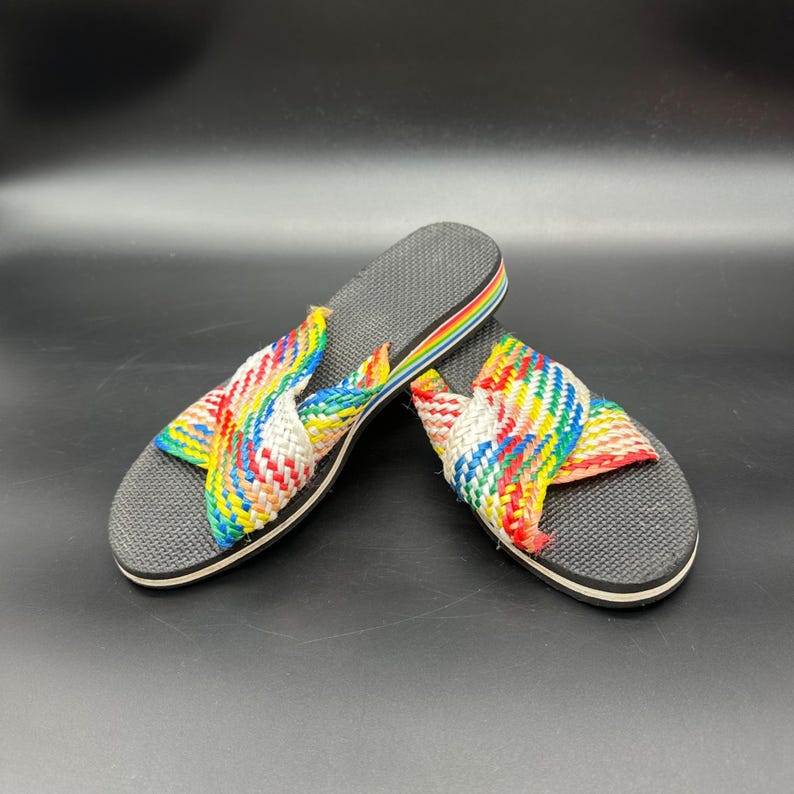 May include: A pair of colorful woven sandals. The sandals have a gray sole with a rainbow stripe along the edge. The straps are woven with red, yellow, blue, and white threads.
