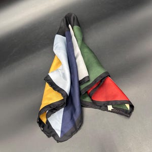 May include: A square silk scarf featuring a color-blocked design with distinct sections of mustard yellow, light blue, navy blue, white, forest green, and red. The scarf has a thin black border along all edges and a smooth, reflective finish.