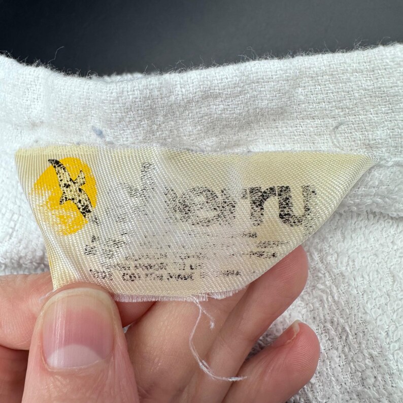 Op de afbeelding: Een close-up van een label met het woord "Veru" in zwarte letters. Het label heeft een geel en zwart vogel logo. Het label staat op een witte, getextureerde stof.