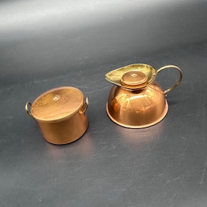 Puede incluir: Dos artículos de cocina de cobre: una olla pequeña con tapa y una crema con asa y boquilla de latón. La olla tiene dos asas pequeñas. La crema tiene un cuerpo redondo y una tapa de latón.