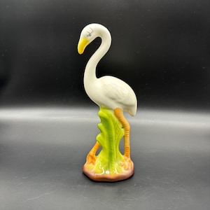 Puede incluir: Una figura de cerámica de un flamenco blanco con pico amarillo y detalles negros en los ojos. El ave se apoya sobre una base verde y marrón que recuerda al follaje. El flamenco tiene patas naranjas y un cuello largo y curvado, acabado brillante.
