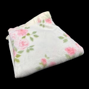 May include: Folded white blanket with a floral pattern of pink roses and green leaves. The blanket has a soft, plush texture and a light yellow satin trim along the edge. The roses are scattered across the surface.