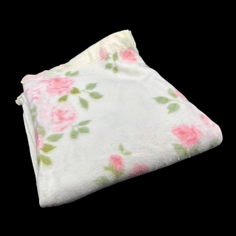 May include: Folded white blanket with a floral pattern of pink roses and green leaves. The blanket has a soft, plush texture and a light yellow satin trim along the edge. The roses are scattered across the surface.
