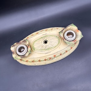 May include: An oval-shaped metal ceiling light fixture base with a cream-colored finish, featuring green and orange floral accents. The fixture includes two white ceramic light bulb sockets surrounded by decorative petal-shaped metal edges.