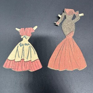 Puede incluir: Dos vestidos de mu&ntilde;ecas de papel vintage. Uno es un vestido rosa y crema con una falda amplia y un top a juego. El otro es un vestido largo color coral con un corpi&ntilde;o ajustado y una falda acampanada.
