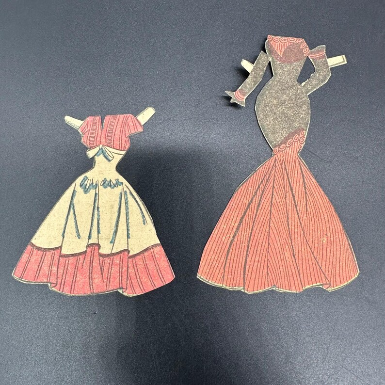 Puede incluir: Dos vestidos de mu&ntilde;ecas de papel vintage. Uno es un vestido rosa y crema con una falda amplia y un top a juego. El otro es un vestido largo color coral con un corpi&ntilde;o ajustado y una falda acampanada.