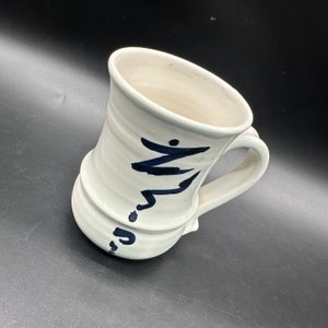May include: White ceramic mug with a black hand-painted design. The design is abstract and features a series of lines and curves.