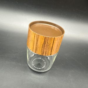 Puede incluir: Un tarro de vidrio transparente con una tapa marrón y una banda con diseño de vetas de madera alrededor de la parte superior. El tarro tiene texto en la parte inferior. El tarro está sobre una superficie oscura.