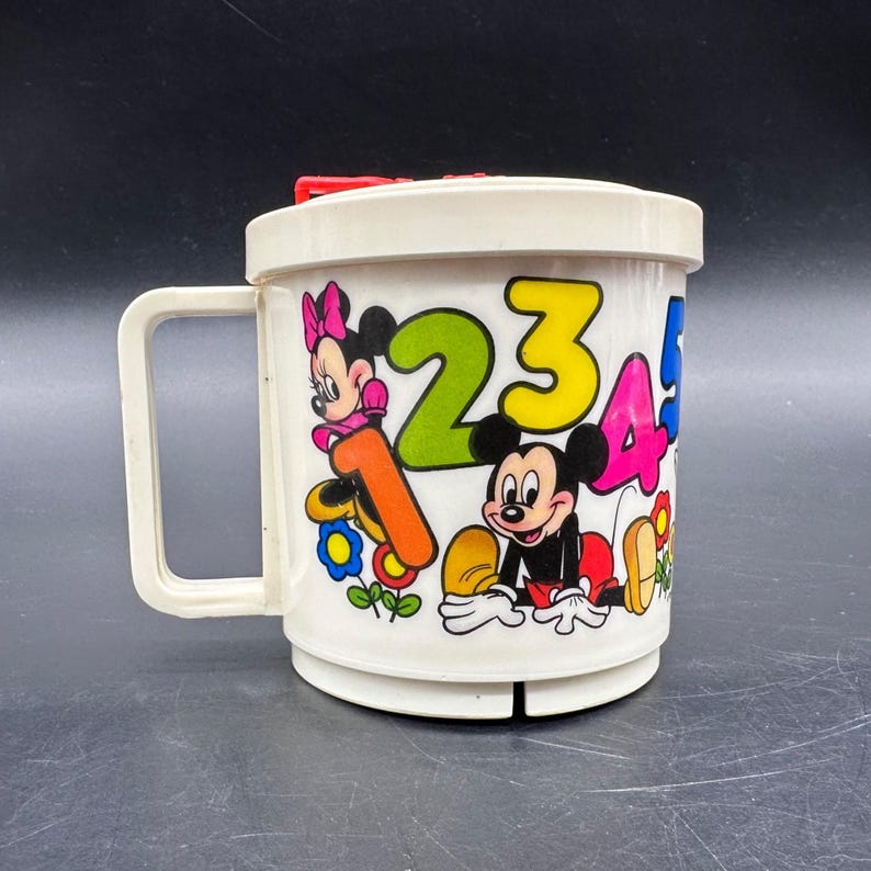 Puede incluir: Taza de pl&aacute;stico blanco vintage con Mickey y Minnie Mouse, con n&uacute;meros coloridos 1, 2, 3, 4 y 5. La taza tiene un asa y una tapa roja. El dise&ntilde;o incluye flores y es probablemente un art&iacute;culo para ni&ntilde;os.