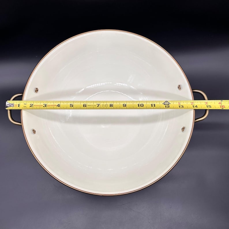 Op de afbeelding: Een ronde, cr&egrave;mekleurige ge&euml;mailleerde pan met twee bronskleurige metalen handgrepen aan weerszijden. Een geel meetlint is over het midden gespannen en toont aan dat de binnendiameter van de pan ongeveer 39,4 cm breed is.
