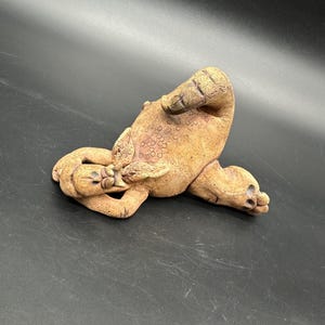May include: A ceramic sculpture of a brown, stylized creature with a textured surface. The creature is lying on its back with its legs in the air.
