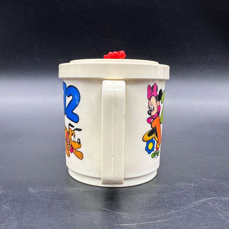 Puede incluir: Taza de pl&aacute;stico blanquecina con personajes de dibujos animados. La taza tiene un asa y una tapa roja. El n&uacute;mero 12 en azul es visible, as&iacute; como Pluto y Minnie Mouse. La taza es probablemente vintage.