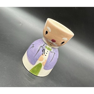Peut inclure: Un coquetier en céramique en forme de Père Noël. Le coquetier a un visage brun clair avec des yeux noirs, un nez rouge et une barbe blanche. Le corps est violet avec un motif vert et blanc.