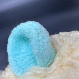 Peut inclure: Gros plan sur une oreille de jouet en peluche. L'oreille est bleu clair et a une texture douce et floue. La fourrure environnante est blanc cass&eacute;. Le fond est de couleur fonc&eacute;e.