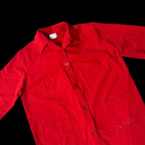 May include: A bright red button-down shirt with a collar and two front pockets. The long-sleeved shirt has a classic design and is made of a smooth fabric. The shirt has a small tag on the collar.
