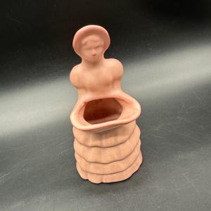 Puede incluir: Un jarrón de cerámica vintage con forma de mujer, pintado en un color melocotón apagado. El jarrón presenta un rostro detallado, un sombrero y un diseño de falda en capas. La abertura tiene forma ovalada.