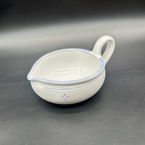 May include: A white ceramic gravy boat with a handle and a pouring spout. The boat is decorated with light blue trim and small blue dot clusters. The handle is arched, and the boat has a rounded shape.