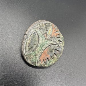 May include: A handcrafted, oval-shaped ceramic pendant with an abstract design. The pendant features a central green and orange motif, surrounded by a textured, gray-brown border. The design includes yellow accents.