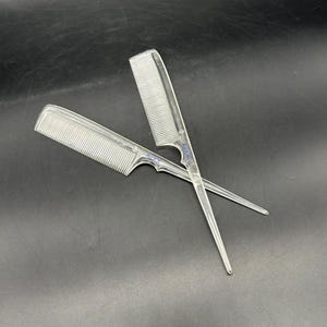 May include: Two clear plastic combs are crossed against a dark background. The combs have fine teeth and a handle with a pointed end. The brand name "AJAX" is printed on the handle.