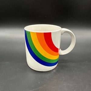 May include: White ceramic mug with a rainbow design. The rainbow is a curved arc of red, orange, yellow, green, blue, and indigo.