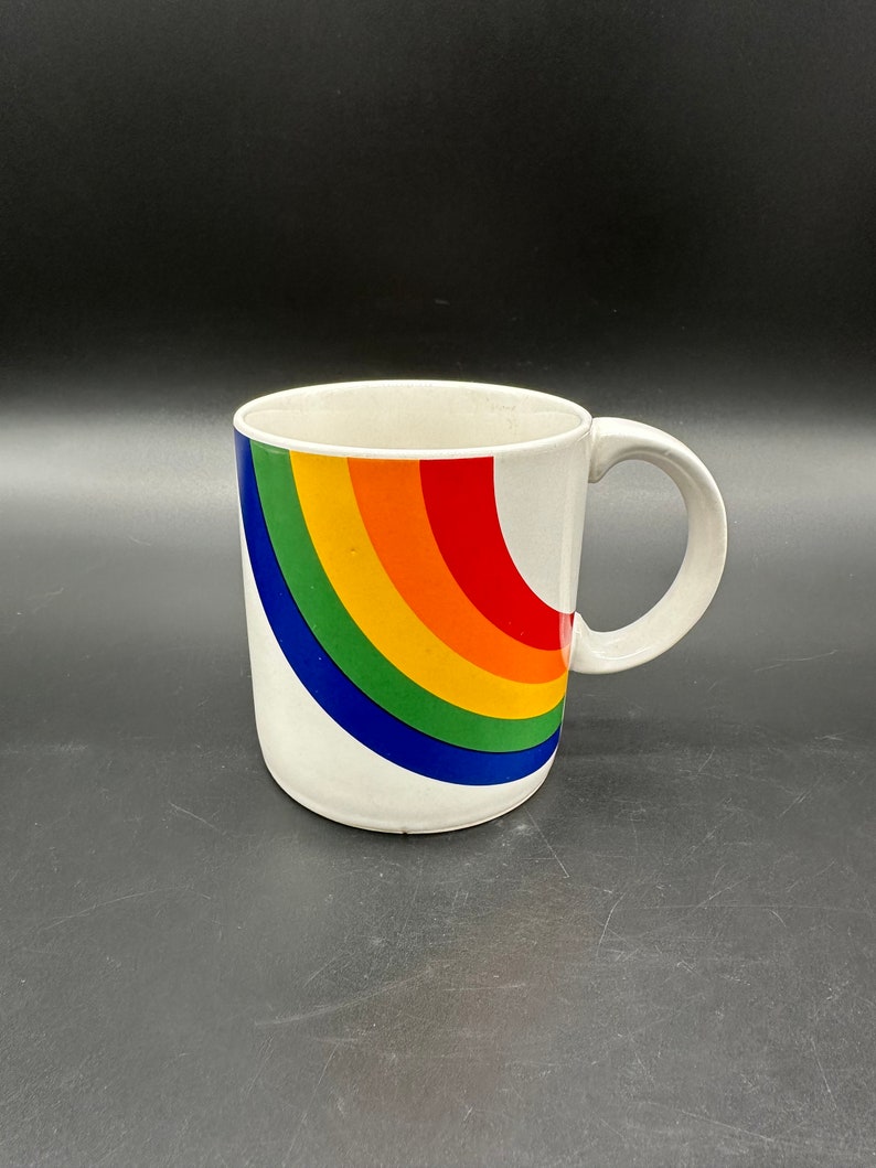 May include: White ceramic mug with a rainbow design. The rainbow is a curved arc of red, orange, yellow, green, blue, and indigo.