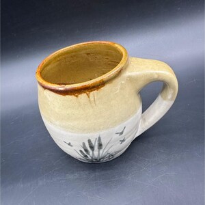 May include: A handmade ceramic mug with a unique design. The mug features a cream-colored upper half and a white lower half, with a hand-painted design of cattails and birds. The rim is a dark brown color, and the handle is curved.
