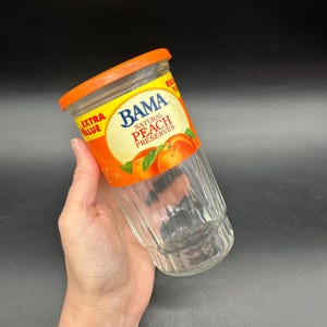 May include: A clear glass jar with an orange lid and a label that reads "BAMA Natural Peach Preserves." The label features peaches and green leaves. The jar is held by a hand against a dark background.