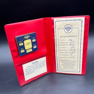May include: Open red case with a blue and gold gem certificate. The certificate displays a topaz gemstone with a carat weight of 1.83. The case also contains a gem data certificate with details about the topaz.