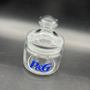 Puede incluir: Un tarro de cristal transparente con tapa y pomo. El tarro tiene un logotipo azul "P&G". El diseño es clásico y el tarro probablemente se utiliza para el almacenamiento.