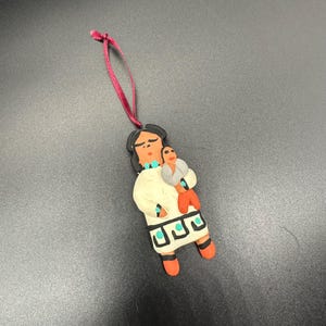 Puede incluir: Un adorno de cerámica hecho a mano que representa a una mujer sosteniendo a un niño. La mujer lleva un vestido blanco con detalles turquesa y el niño lleva un vestido rojo. El adorno está sujeto a una cinta roja.